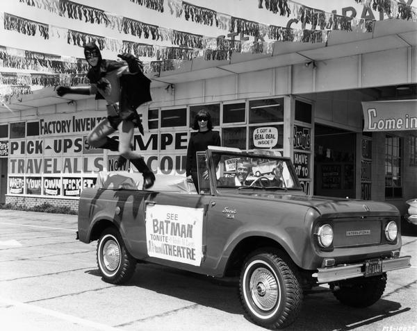 Man dressed in costume as Batman is leaping from the back of an International Scout pickup as part of a promotion for the Norwalk Theatre. The Scout is parked outside an International motor truck dealership. A woman in sunglasses and dark dress is standing up in the truck near a man in the driver's seat.