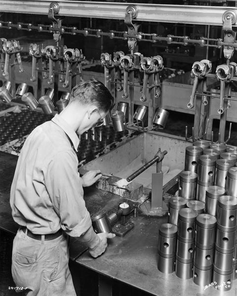 Worker inspecting pistons at International Harvester's Indianapolis Motor Truck Engine Works. Original caption reads: "An inspection operation on the wrist pin holder and lock ring groove on piston. Pistons are inspected 100 percent on this operation. Pin holes must be held to .0001 in. Note power conveyor on which the pistons are hung to be carried along with the connecting-rod assemblies to the final piston and connecting-rod assembly."