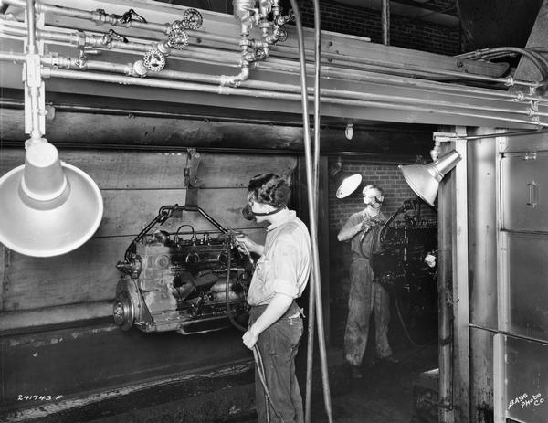 Factory workers spray-painting truck engine blocks in a paint room of International Harvester's Indianapolis Truck Engine Works. Original caption reads: "DeVilbis water-wash type paint booths used for painting all engines. This is one of the most modern pieces of equipment in use for this purpose. It will be noted that the booths are staggered so that when an operator is spraying one side the other operator is out of direct line with the spray. All over-spray is removed from paint booths by a water-wash system that, by means of suction fans, draws the air through a veil of running water. Thus, the operators breathe only thoroughly cleaned air."
