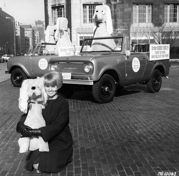 Woman posing with "Cuddly Dudley" stuffed animals (dogs) and International Scout pickups as part of a  promotional campaign. A sign on each Scout reads: "Order Cuddly Dudley from the man who calls at your home."