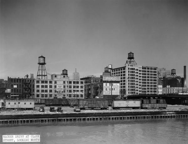 Chicago's Wacker Drive at Clark Street, looking north. Buildings, water towers and railroad boxcars are along the Chicago River waterfront, as well as a Coca-Cola billboard and the back of The Hotel Howard.