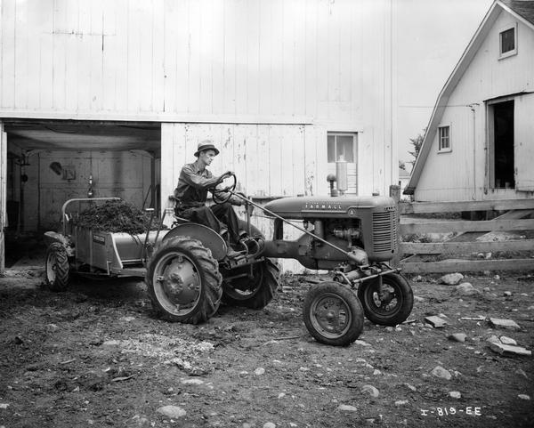 Farmer pulling a loaded manure spreader out of a barn with a Farmall A tractor.