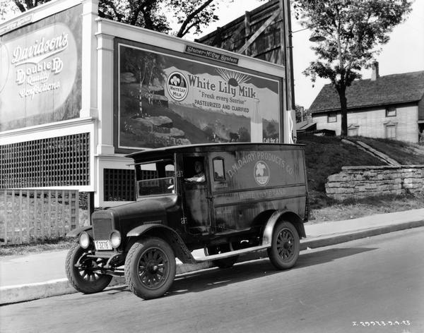 View across street towards a man in the driver's seat of an International Special Delivery dairy truck parked in front of billboards. The International Special Delivery dairy truck is owned by D.M. Dairy Products Co. In the background are billboards for White Lily Milk, a product of D.M. Dairy Products Co., and Davidson's Confections.