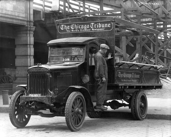 The male driver of the International Harvester Model "63" motor truck, that has been converted for the delivery service of the "Chicago Tribune" Newspaper, is standing on the outside foot runner and looking to the rear. The sign on the truck reads: "The World's Greatest Newspaper." Behind the truck is a large wooden scaffolding structure.