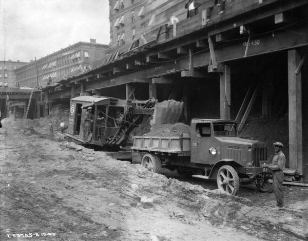 Workers are removing dirt for the construction of the New York City subway with a Bucyrus shovel loader and an International truck. The workers are below street level, and buildings rise above them in the background.