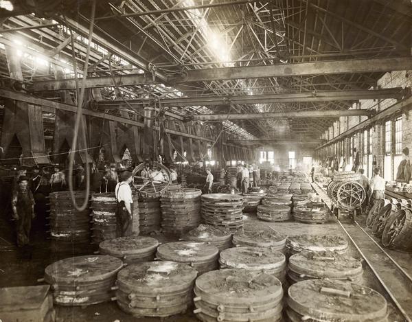 The original caption reads as follows: "Harvesting machines are made largely of iron and steel, consequently foundries, blacksmith shops, machine shops and forge rooms predominate. This picture is taken in one of the numerous grey iron foundries where parts of harvesting machines are made. It shows a section devoted to casting mower wheels. This foundry produces over 200,000 of these wheels a year. This particular foundry annually converts 65,000 tons of pig iron into castings, numbering 20,000,000 pieces annually. A thousand men are employed therein, and the best possible type of molding machines and devices are used to make the molds in which the parts are cast. The foundries are equipped for handling the molten metal and castings with the greatest facility and without unnecessary heavy hand lifting."