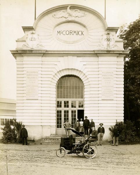 Man sitting in a McCormick Auto-Mower in front of the McCormick Harvesting Machine Company building at the Paris Exposition in France. Several men, a woman, two children and a French police officer are looking on. The Auto-Mower was an experimental gasoline powered mowing machine. This is one of two Auto-Mowers produced by the company. This machine had a two-cylinder engine. The other machine had a single-cylinder engine.