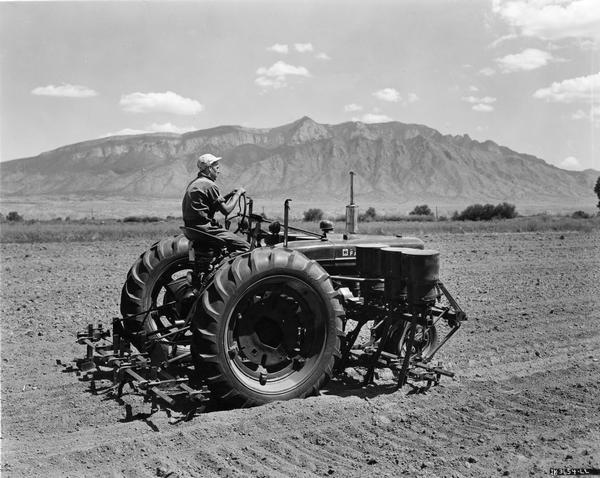Farmer demonstrating 6-row forward-mounted beet and bean cultivator for Farmalls M, MD, or H at the U.S. Soil Conservation Albuquerque Demonstration. Caption reads: "Rows may be 16 to 22 inches apart. It is raised or lowered by Farmall hydraulic Lift-All. Parallel lift for front and rear sections is a feature of this outfit. A 6-row fertilizer attachment is also available."