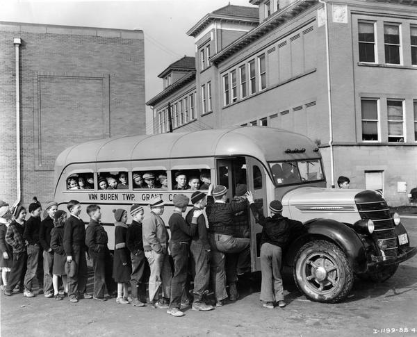 Children line up to board a crowded International D-30 school bus outside a school building. The bus served Van Buren township in Grant County, Indiana. The bus featured a 155-inch wheelbase and a 16-foot Hicks body with a 36-passenger capacity.
