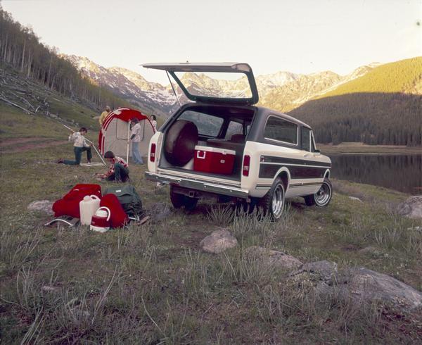 Color advertising photograph of a family setting up a tent in mountain field near a small lake. An International Traveler pickup with simulated vinyl roof, wood grain side applique panels, side moldings, and trailer towing package is featured.