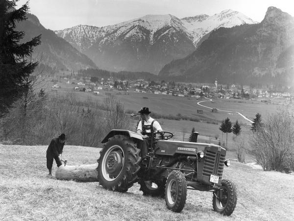 German farmers pulling a wooden log with a McCormick D-435 tractor against a mountainous backdrop in Alpine country.