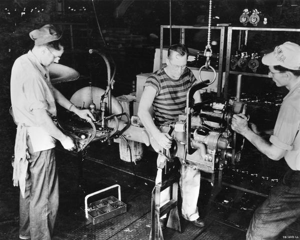 Two factory workers are attaching a Farmall Cub engine to the clutch housing, while a third worker is attaching brake pedals. Taken on the assembly line at International Harvester's Louisville Works.