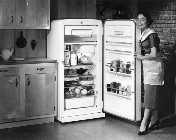 Advertising photograph of a woman wearing an apron over a dress holding the door of an International Harvester refrigerator open.