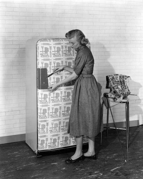Advertising photograph of a woman putting a fabric covering on an International Harvester refrigerator. The refrigerator was designed to accomodate custom designs for interior decoration.