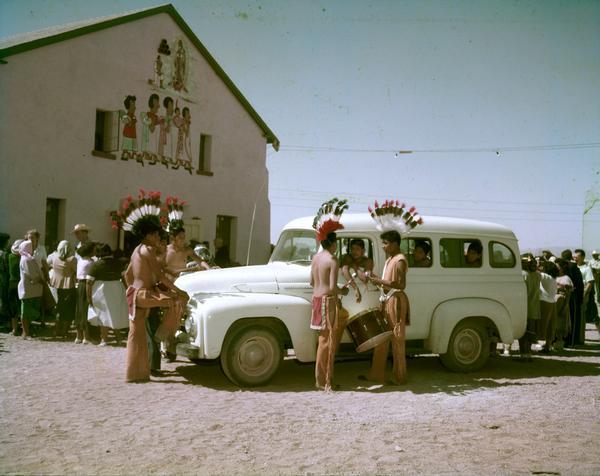 Color photograph of young Native-American men dressed in traditional clothing standing around an International R-Line truck. The young men are with a small crowd of men and women gathered outside what appears to be a meeting hall or building.