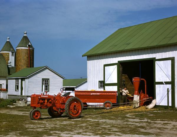 Farmer standing in open barn door shelling corn with belt-driven McCormick hammer mill run by a Farmall C tractor.