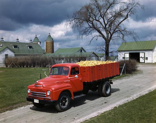 Slightly elevated view of an International L-160 or L-180 truck hauling corn cobs, with a barn and other farm buildings in the background.