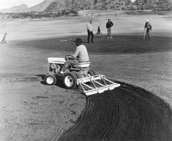 Groundskeeper raking a golf course sand trap with an International Cub Cadet tractor. A group of men are playing golf in the background. The photograph was submitted to International Harvester's Patent Department.