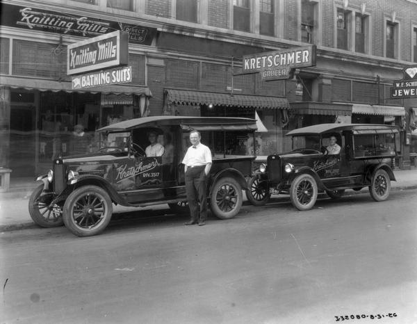 Two drivers and a grocer posing with International delivery trucks outside Kretschmer's grocery store. Other businesses on the street include the Associated Knitting Mills Company and a jewelry store.