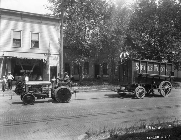Workers pulling a Sells Floto Circus wagon with a McCormick-Deering industrial tractor along the street of a town. In the background, children and adults are looking on from a meat market storefront.