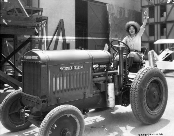Young woman in "country" or riding costume waving from the seat of a McCormick-Deering industrial tractor. The woman may be an actress and appears to be on or outside a studio set. A small airplane is in the background.