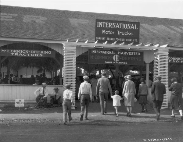 Adults and children approaching the entrance of International Harvester's pavilion at the Iowa State Fair. Signs on the building advertise "International Motor Trucks" and "McCormick-Deering Farm Machines."