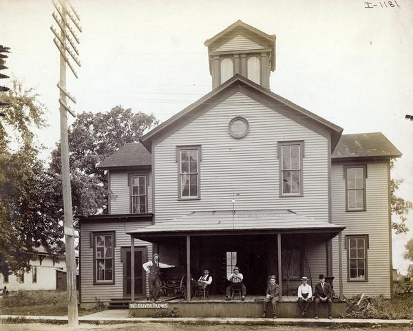 A group of men sitting and standing on the front porch of a building. The building appears to be a former schoolhouse and may be an International Harvester dealership. The porch has a sign for Oliver Plows, a wagon and a feed grinder.