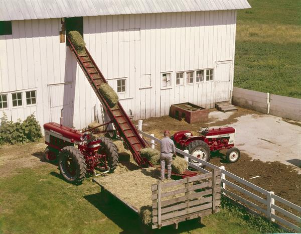 Elevated view of a farmer using a hay loader (elevator) to transport hay bales into a barn loft. A Farmall 504 utility tractor and a Farmall 460 tractor are parked in the foreground.