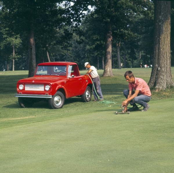 Color photograph of two groundskeepers preparing to water a golf course green. The men are using a 1972 Scout 4x4 pickup for transportation and hauling equipment.