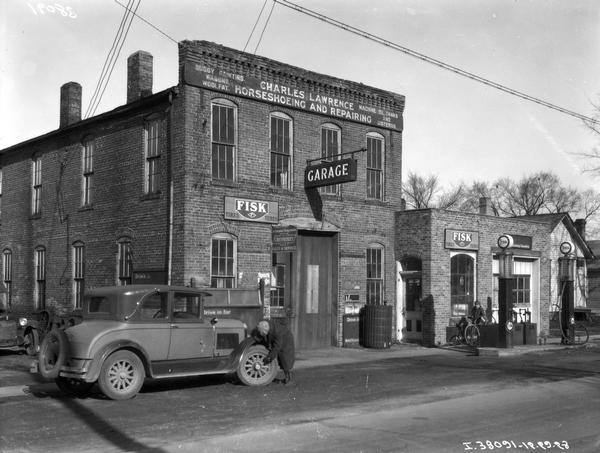 View across street towards a man checking an automobile tire in front of a service station and garage owned by Charles Lawrence. A boy is standing with his bicycle in the background.