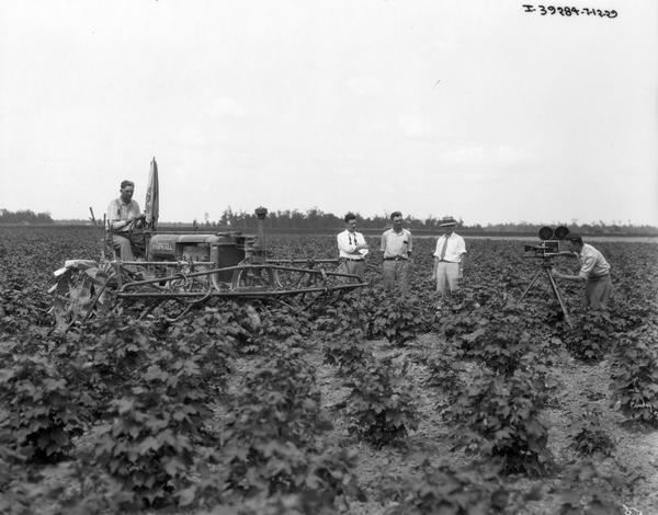 Cameraman filming a farmer on a McCormick-Deering Farmall Regular tractor with attached cultivator as three men look on.