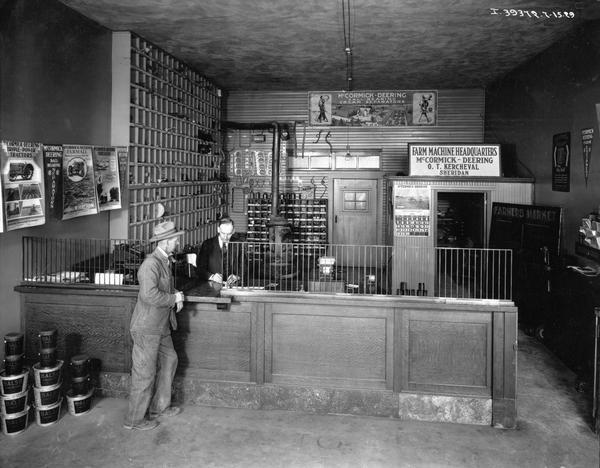 Customer standing at the parts desk of an International Harvester agricultural equipment dealership owned by O.T. Kercheval of Sheridan, Indiana. Advertising posters and parts are hanging on the walls.