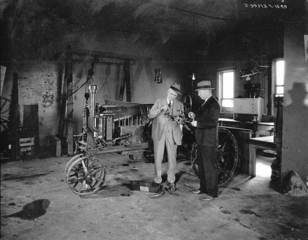 Two men examining parts from a disassembled Farmall Regular tractor in the repair shop of a dealership.