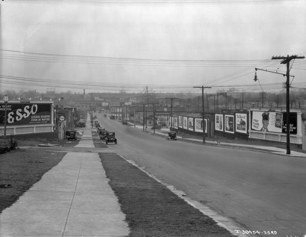 Sherwood Avenue lined with parked cars and billboards advertising "Esso," "Miller Bros." restaurant, "Wrigley's" chewing gum, "Koester's Honey Bread" and other products.