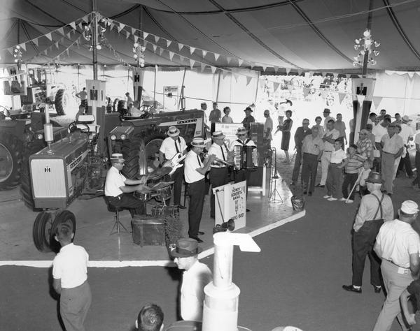 The 'Howdy' Roberts Band performing inside an International Harvester exhibit at the Iowa State Fair. Band members are playing a clarinet, trumpet, accordian, electric bass and drums. Two Farmall 656 tractors are in the background.