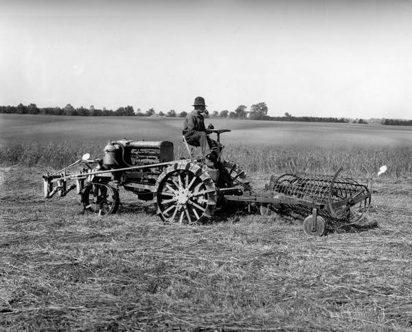 Man operating an experimental(?) reverse drive tractor with mounted hay rake in a field.