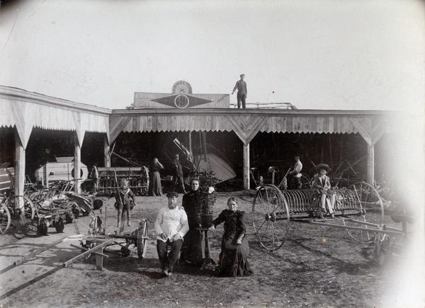 Men, women and children posing among agricultural machinery outside an International Harvester dealership in Russia owned by E.E. Bukin. The machines include a hay rake, mower, grain drill and an Osborne reaper. A potted plant on a stand has been placed between three adults in the foreground. A sign on the building bears the "Osborne" and "Plano" brand names. A man is standing on the roof of the building in the background.