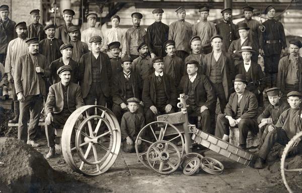 Group of men and young boys posing in the grey iron foundry of International Harvester's Lubertzy Works (factory) in Russia. Pieces of agricultural machinery have been placed in the foreground.