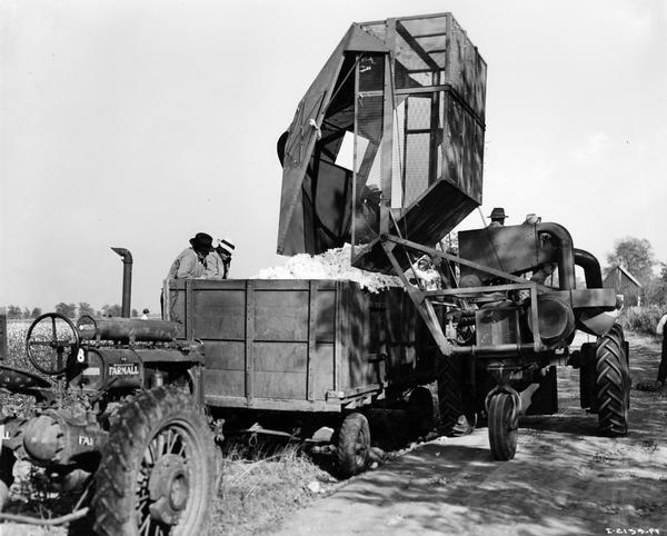 Two men are watching as cotton is unloaded from an experimental International Harvester cotton picker into a wagon. A Farmall tractor is in the foreground.