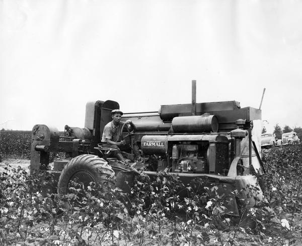 Man operating an experimental International Harvester cotton picker in a cotton field on the Hopson Plantation near Clarksdale, Mississippi.  Two automobiles are parked in the background. Original caption reads: "View showing picker unit side of International Harvester cotton picker. Picker unit is raised or lowered by turning screw near right arm of operator."