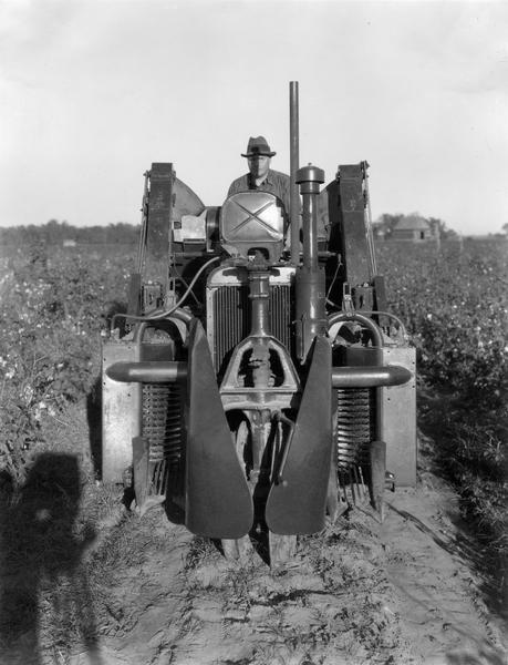 Man operating an experimental 2-row cotton picker built around a Farmall Regular tractor [original caption reads: "Regular Farmall & 2 row cotton picker"]. The photographer's shadow is at the lower left corner.
