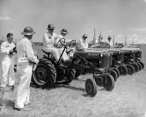Men in jumpsuits and pith helmets on Farmall Cub tractors lined up to demonstrate the tractor and implements. The demonstration was staged in conjunction with a branch meeting.
