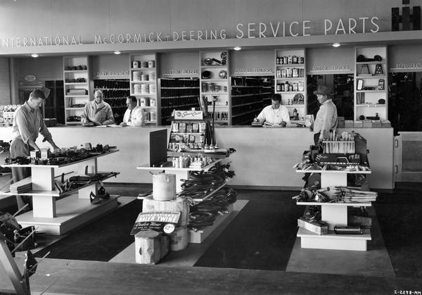 Interior of an International Harvester prototype dealership building owned by Allied Equipment Co. One customer is standing at the service counter and another is looking at parts on the sales floor.