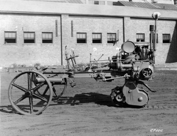 Engineering photograph of an experimental tractor or motor cultivator with a rear crawler track near an International Harvester factory. The original caption reads: "A still further development of the crawler rear end. Note the belt pulley and the third type of power take-off, also the arm projecting rearwardly to provide a hitch for plow and other implements. The three developments shown in prints 29, 30, and 31 indicate considerable activity on the part of the experimental department at this time."