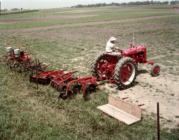 Elevated view of a farmer backing a Farmall Super C tractor up to a Fast-Hitch plow. Other Fast-Hitch implements are nearby including a planter, spring tooth harrow, and disk harrow.