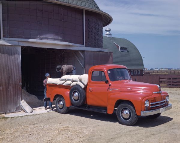 View across barn yard towards two men loading bags into an International L-120 truck. The truck is parked outside a round barn.