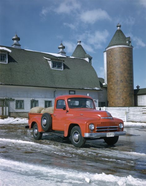 View towards a man sitting in an International pickup truck parked on a snow and ice covered drive outside a barn. The bed of the truck is loaded with filled sacks.