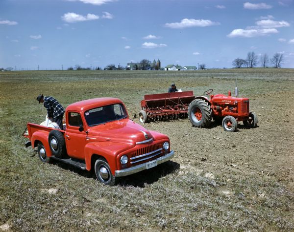 Slightly elevated view of two farmers working in a field with a truck, tractor and grain drill. One man is unloading bags of seed from an International L-120 truck, and the other is loading seed into a grain drill hitched to a McCormick standard W-4 tractor.