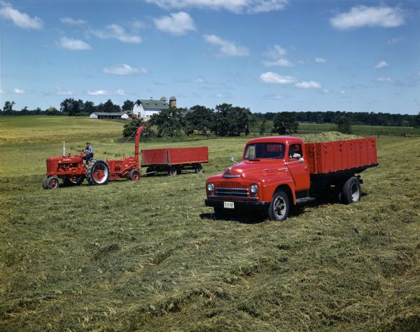 Two farmers are working in a field with a McCormick Farmall M tractor, a 75-P field hay chopper, a trailer and an International truck.