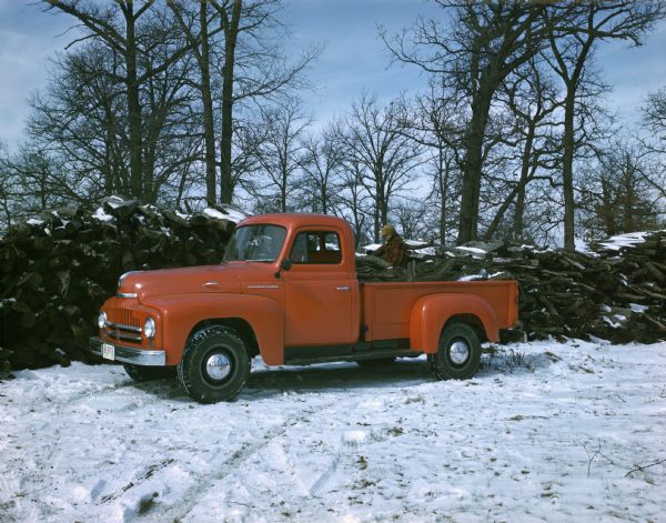 View across snow-covered ground towards a man loading logs for firewood(?) onto an International truck during winter.